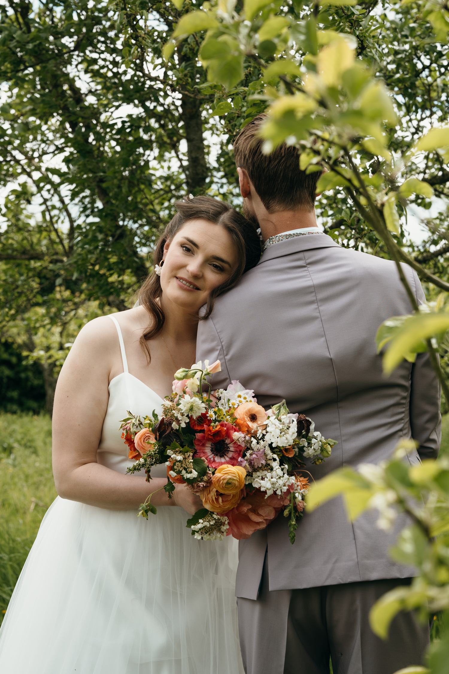 Bride in white dress holds colorful bouquet, leaning on groom in gray suit, surrounded by lush green trees.