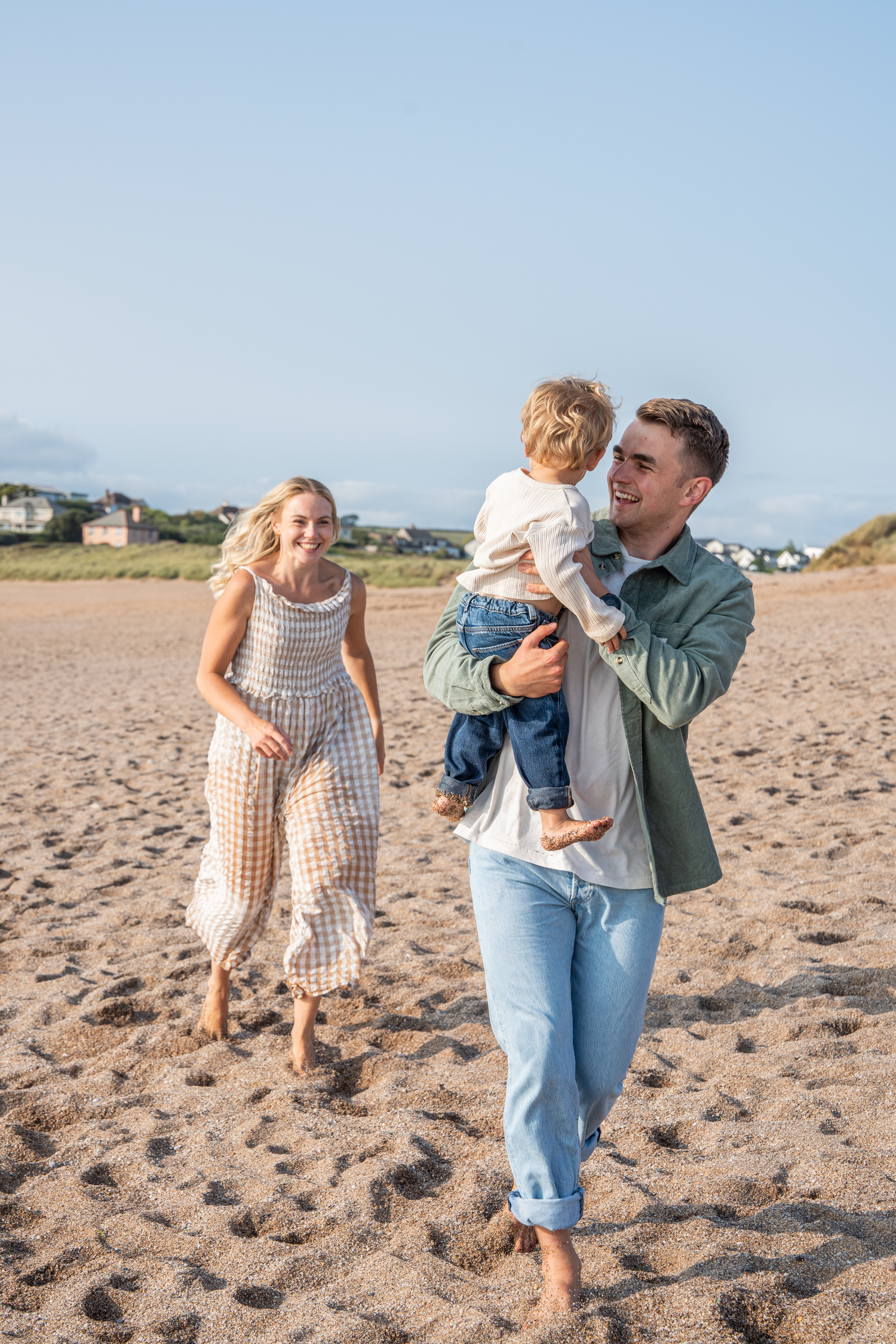 Happy young family walking on a sandy beach, father holding toddler while mother smiles nearby.