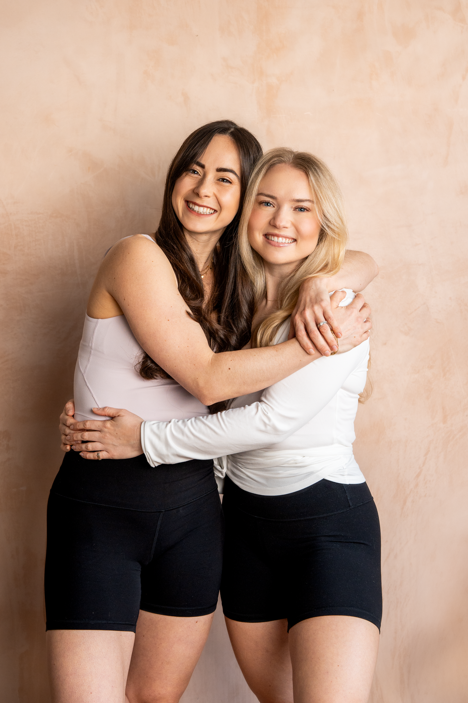 Two smiling young women embracing in matching black shorts against a warm beige studio background.