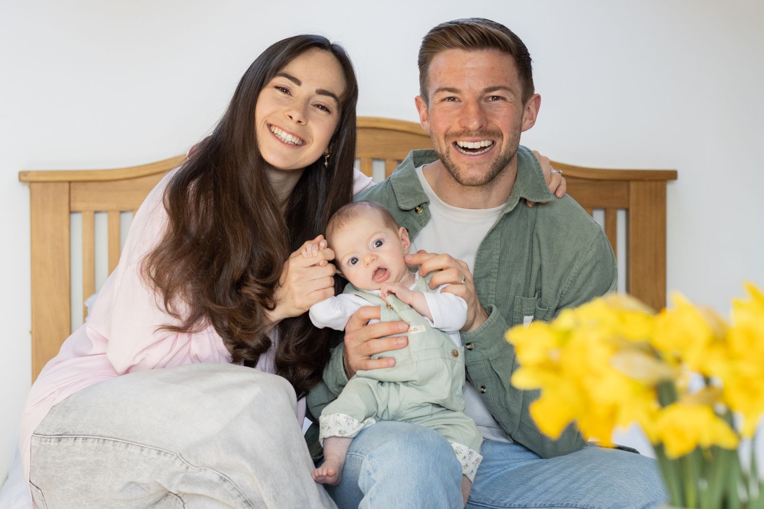 Happy young family with newborn baby sitting on bed with yellow daffodils nearby.