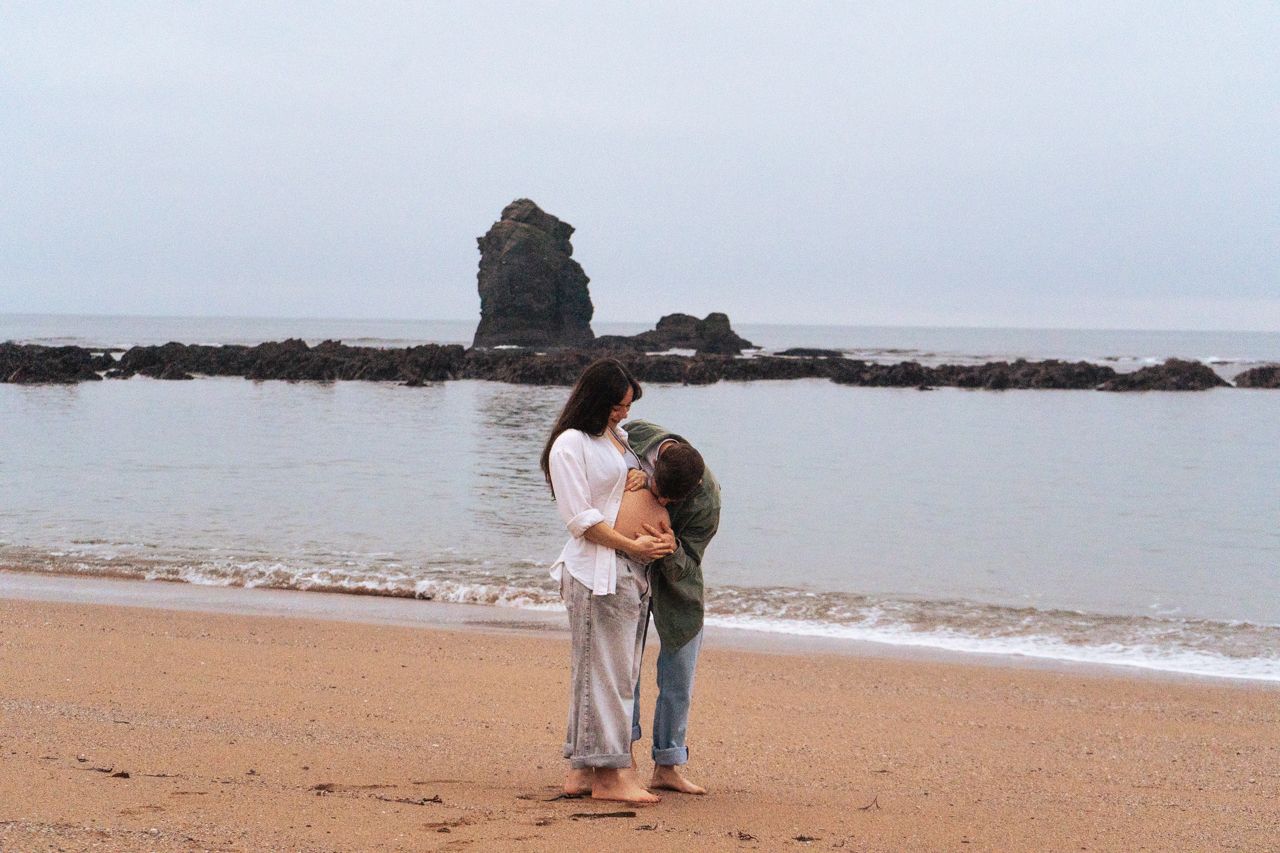 Pregnant couple sharing tender moment on beach with rocky sea stack in background.