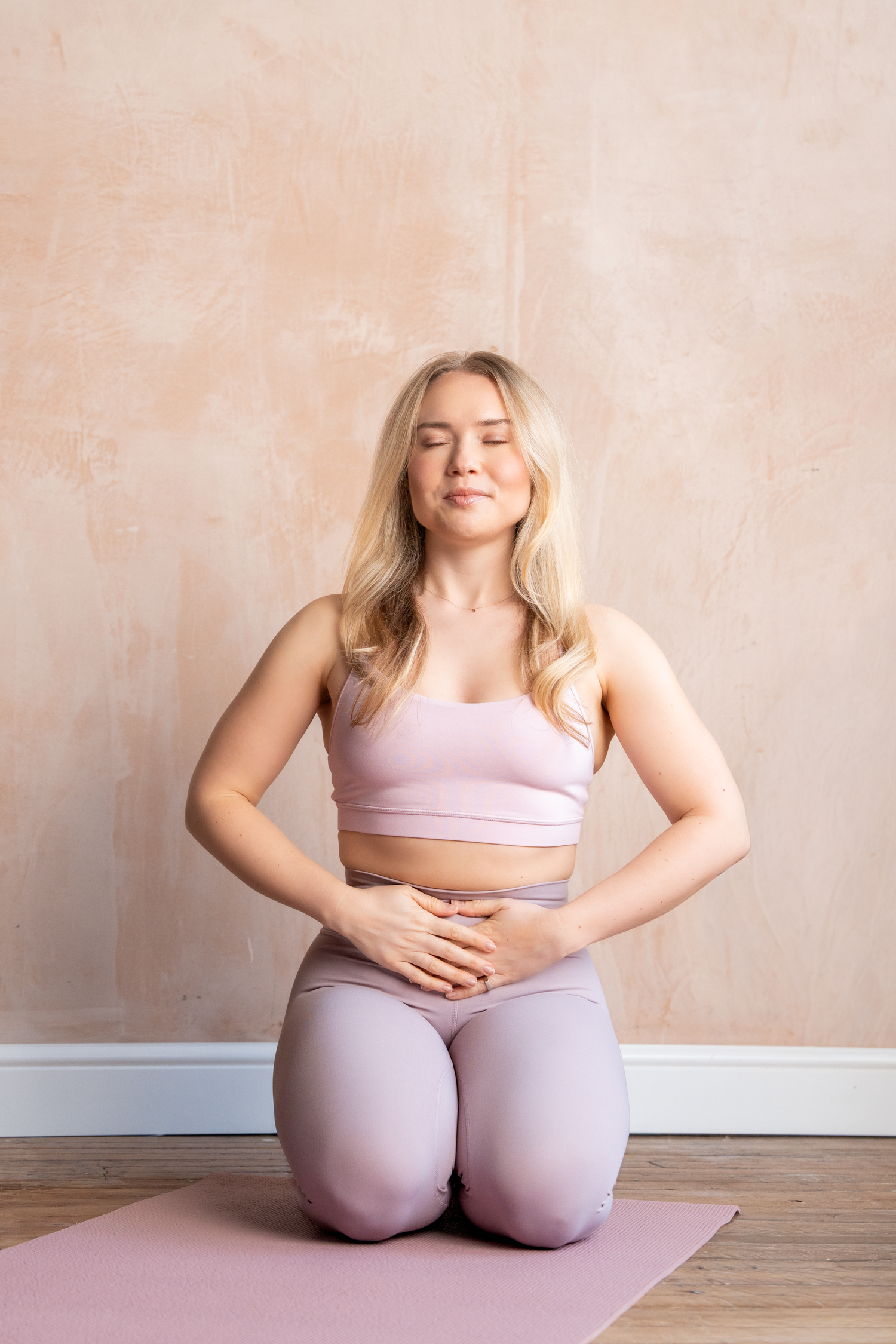 Blonde woman in pink workout set kneeling on yoga mat with eyes closed, hands on stomach.