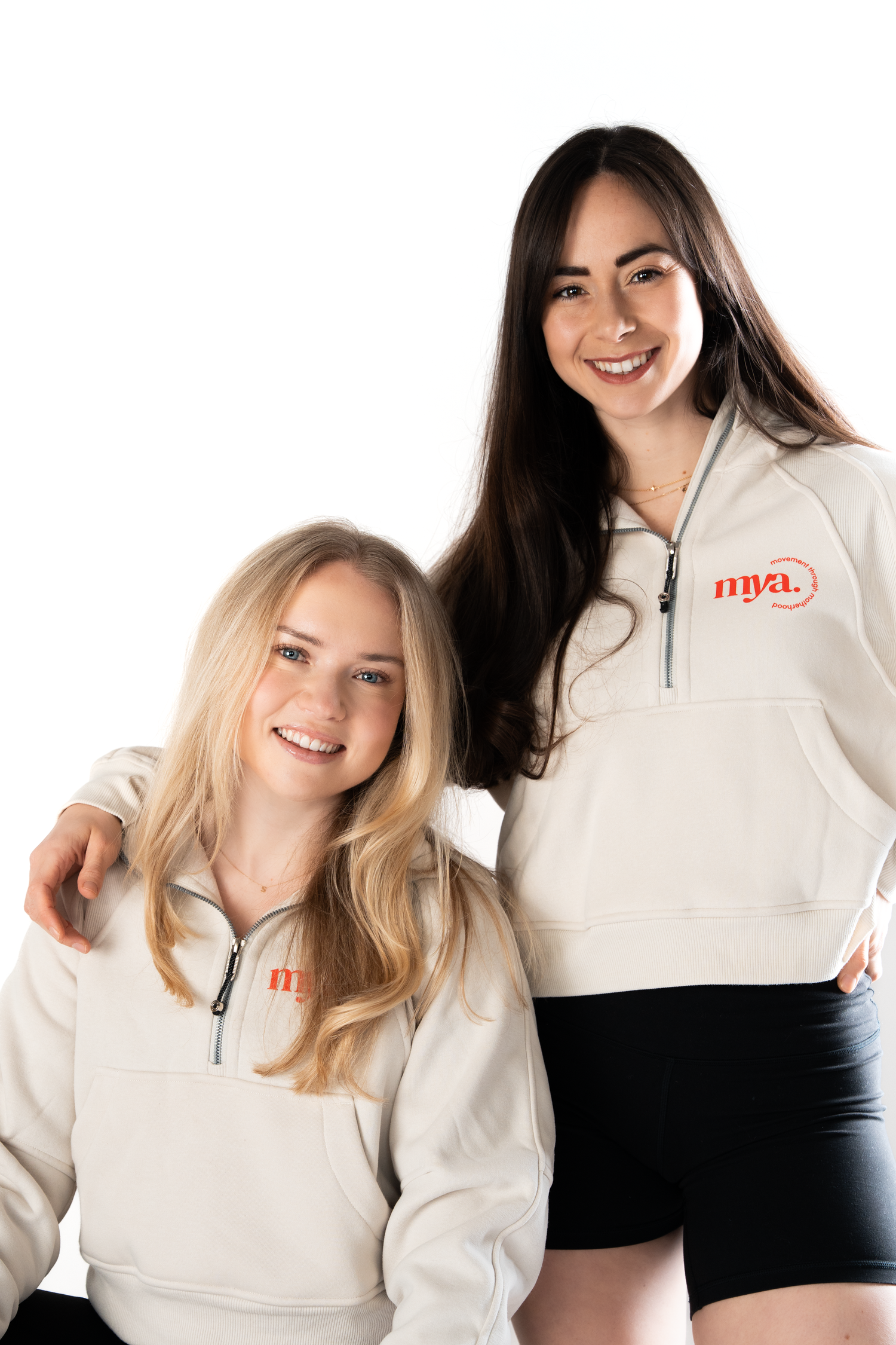 Two smiling young women wearing matching cream NYA branded quarter-zip sweatshirts on white background.
