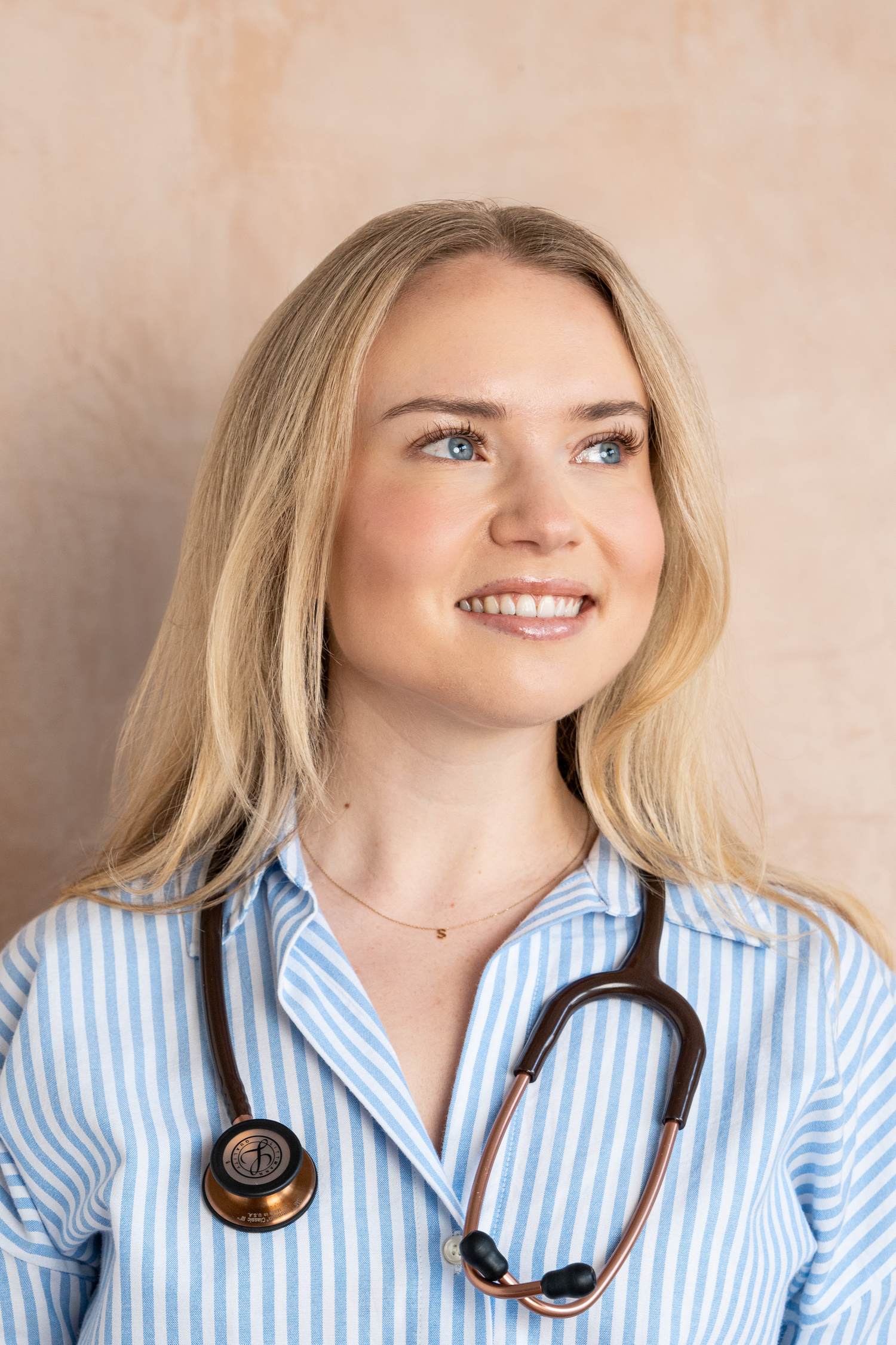 Smiling blonde female doctor wearing a blue striped shirt and stethoscope, looking up confidently.