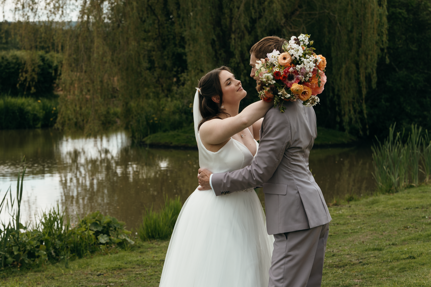 Bride and groom embrace near a pond, bride holding a vibrant orange and white bouquet on their wedding day.