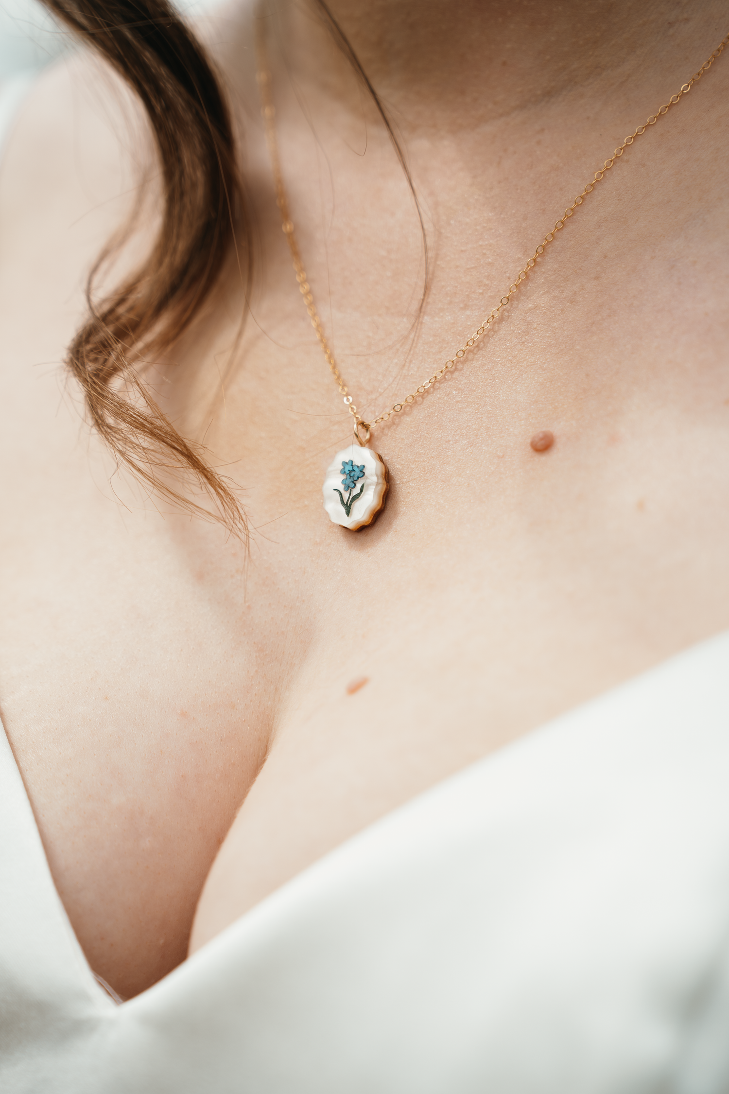 Gold chain necklace with oval white pendant featuring a blue forget-me-not flower, worn by a woman in a white top.