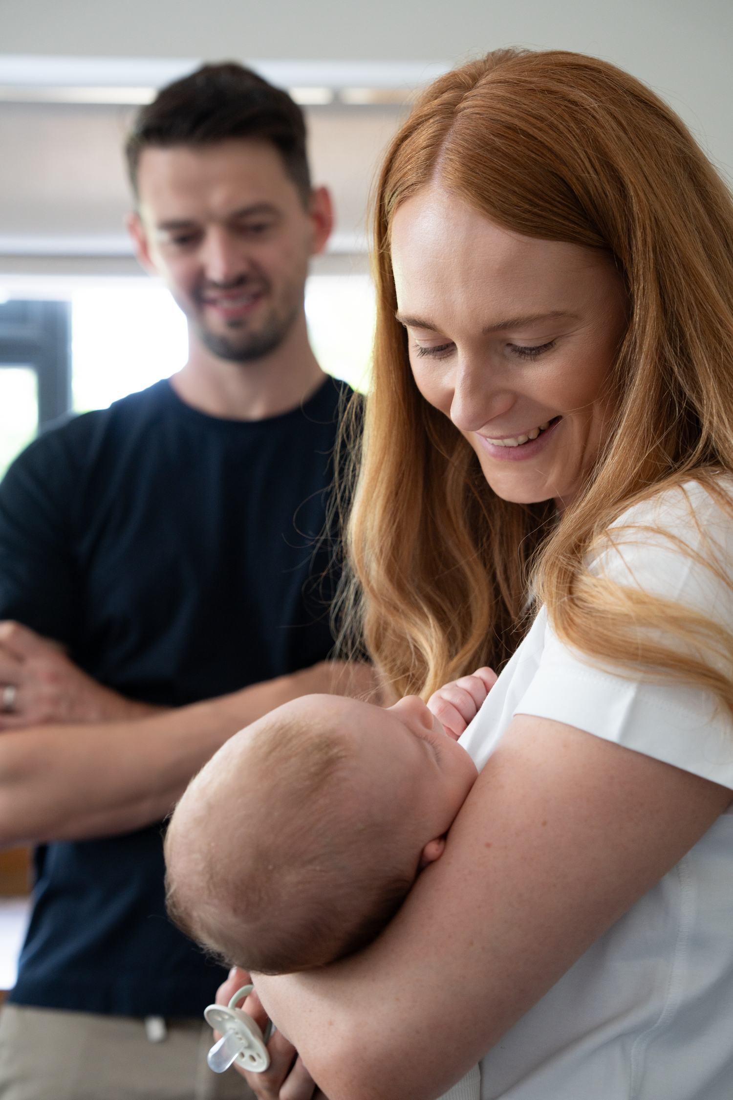 A smiling redhead woman holds a newborn baby with a pacifier while a man in a navy shirt looks on proudly.
