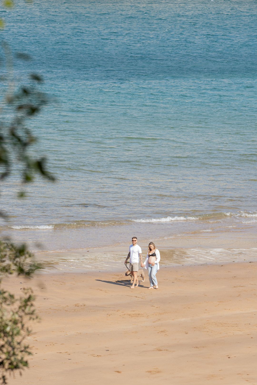Couple walking hand-in-hand along a sunny sandy beach with turquoise ocean waves in the background.