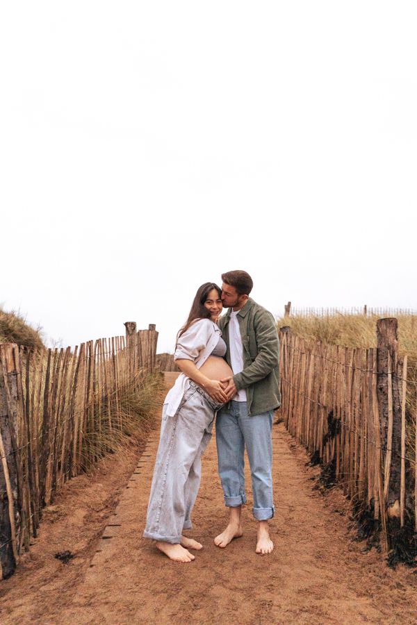 Couple embracing on sandy beach path, man kissing pregnant woman near wooden fence.