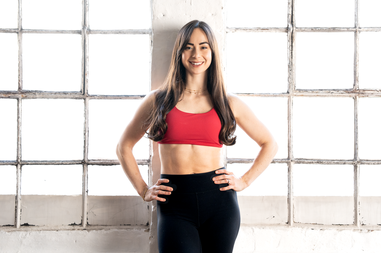 A fit woman in a red sports bra and black leggings smiles with hands on hips in a gym.