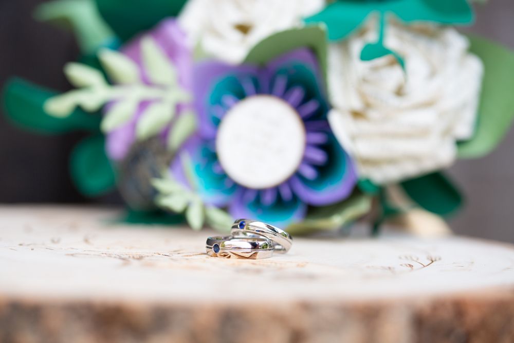 Close-up of wedding rings on a wooden slice with colorful paper flowers in the background.