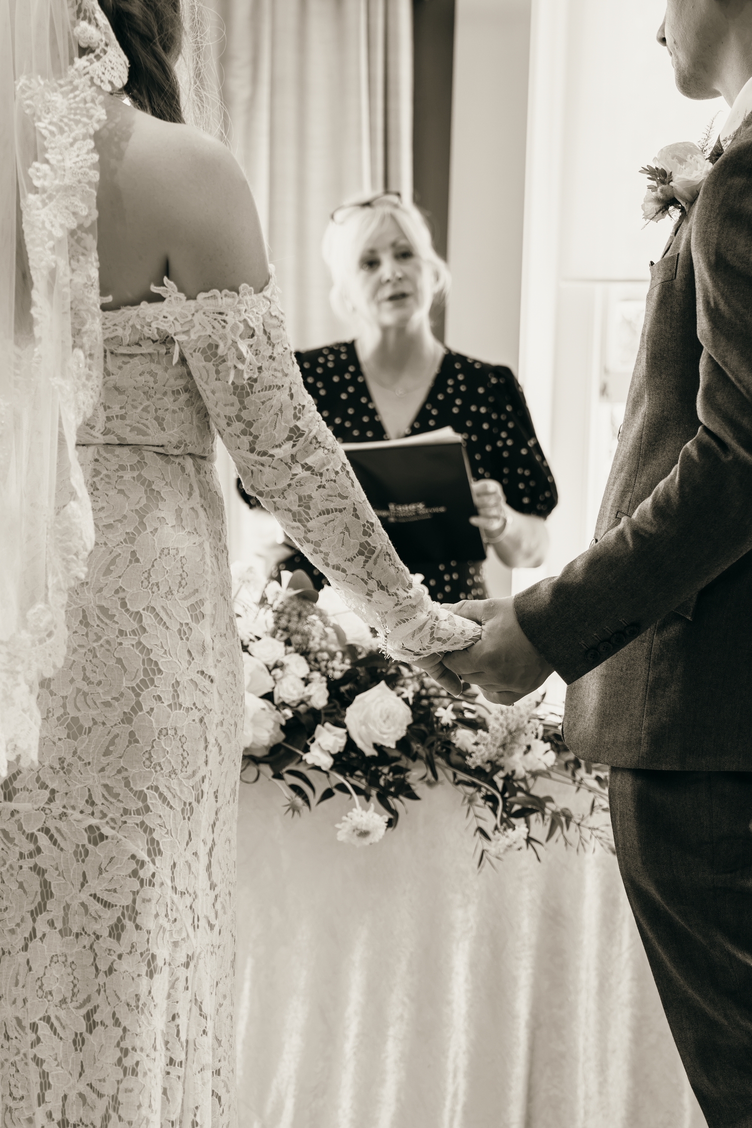 Black and white wedding ceremony photo of bride in lace gown holding hands with groom while officiant reads vows.
