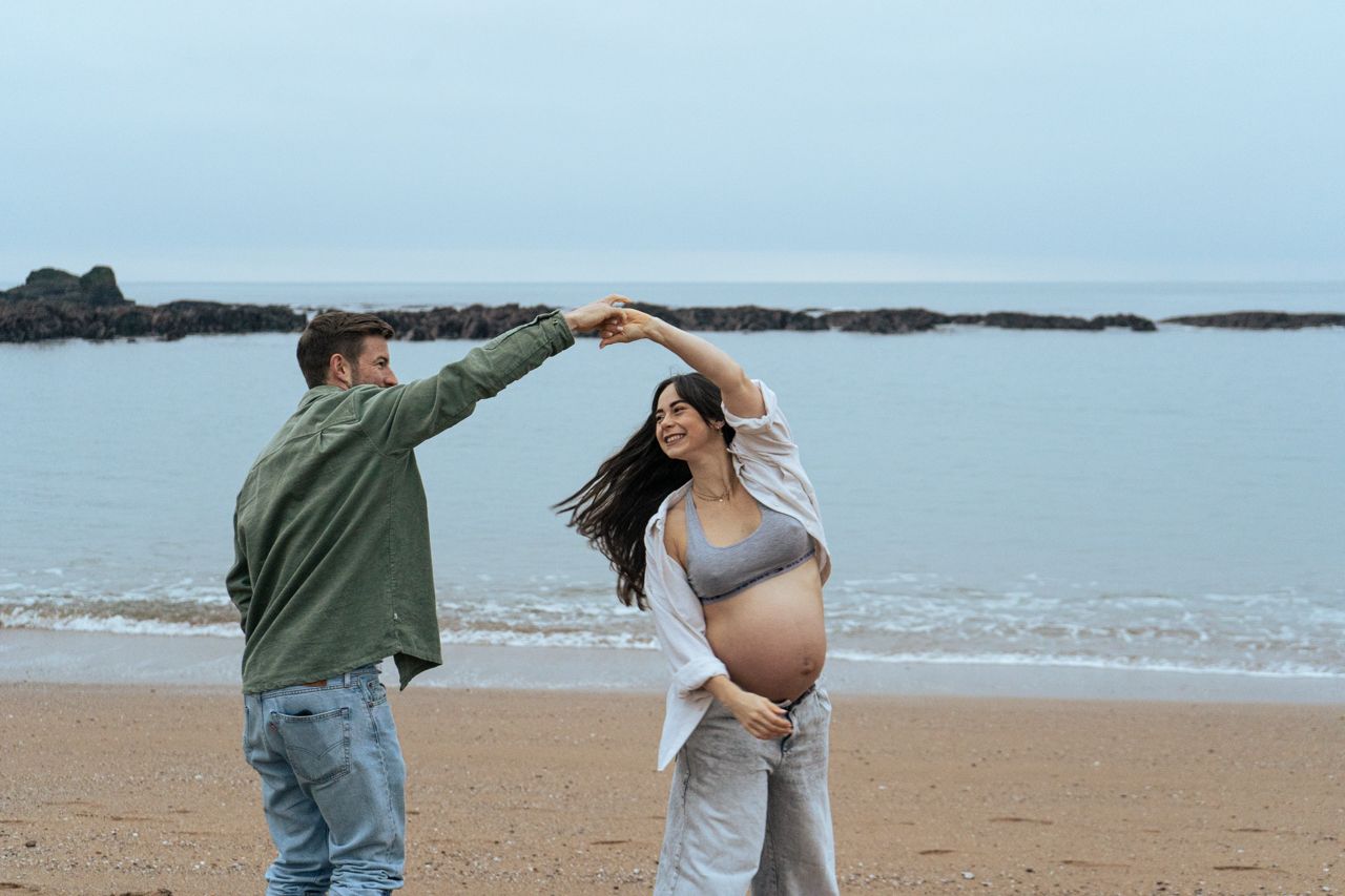 Couple dancing on sandy beach, man twirling pregnant woman as she holds her baby bump.