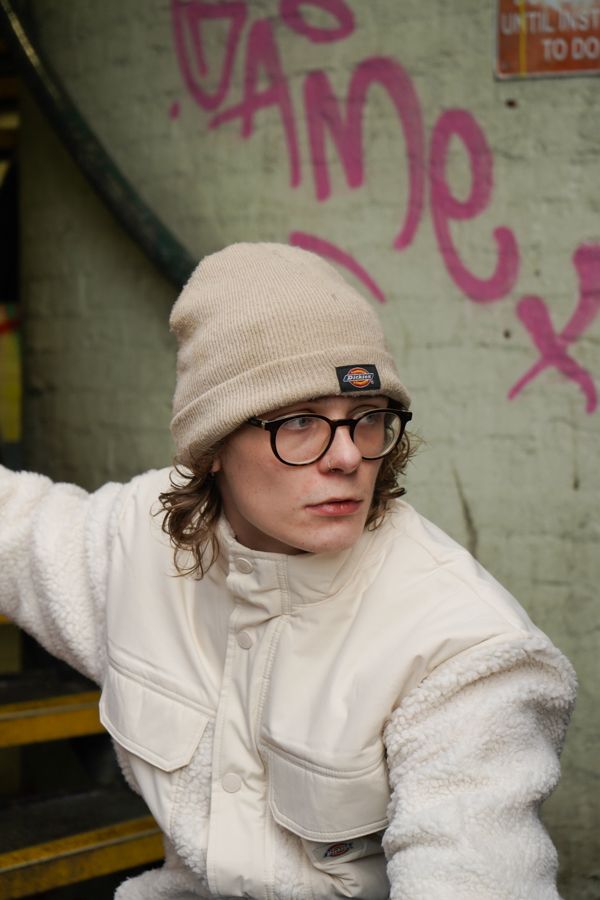 Young person in Dickies beanie and white jacket posing near graffiti wall in urban setting.