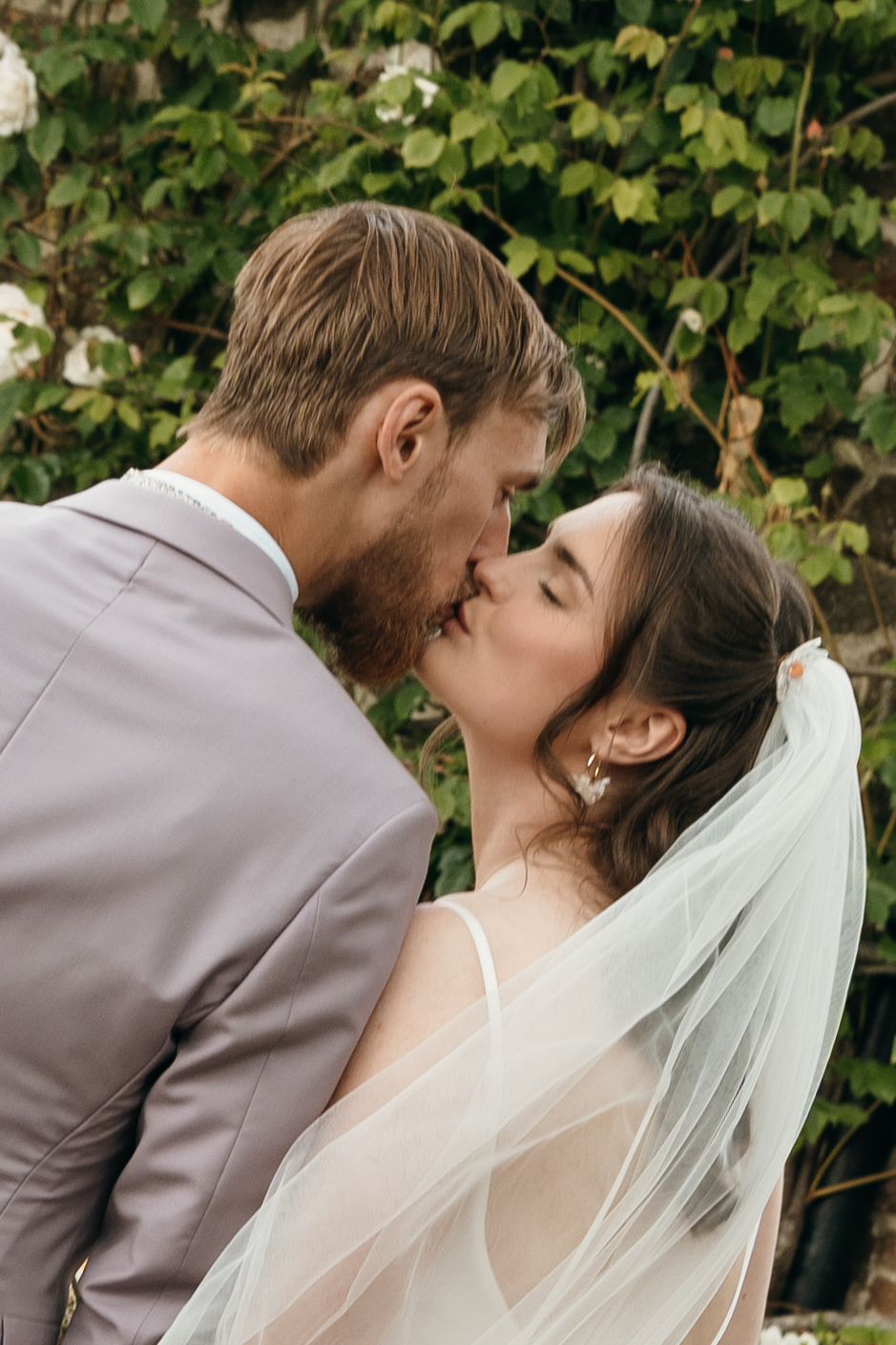 A bride and groom share a romantic kiss outdoors, surrounded by lush green foliage on their wedding day.