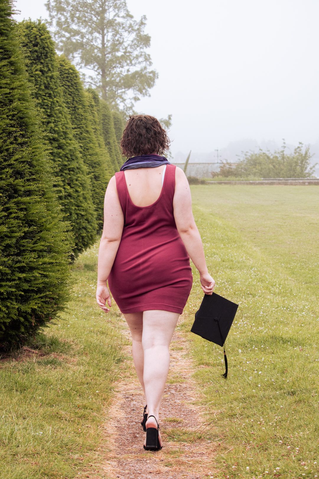A young woman in a red dress holding a graduation cap walks along a garden path lined with tall hedges.