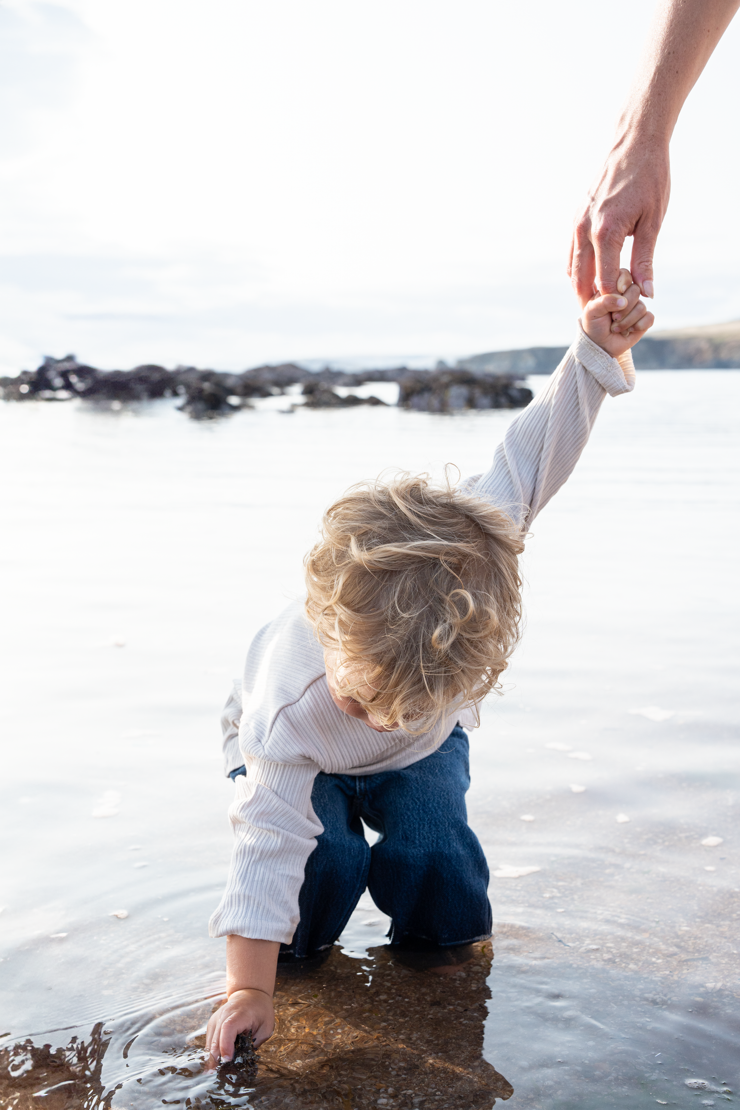 Toddler explores rocky shoreline water while adult hand provides balance and support at the beach.