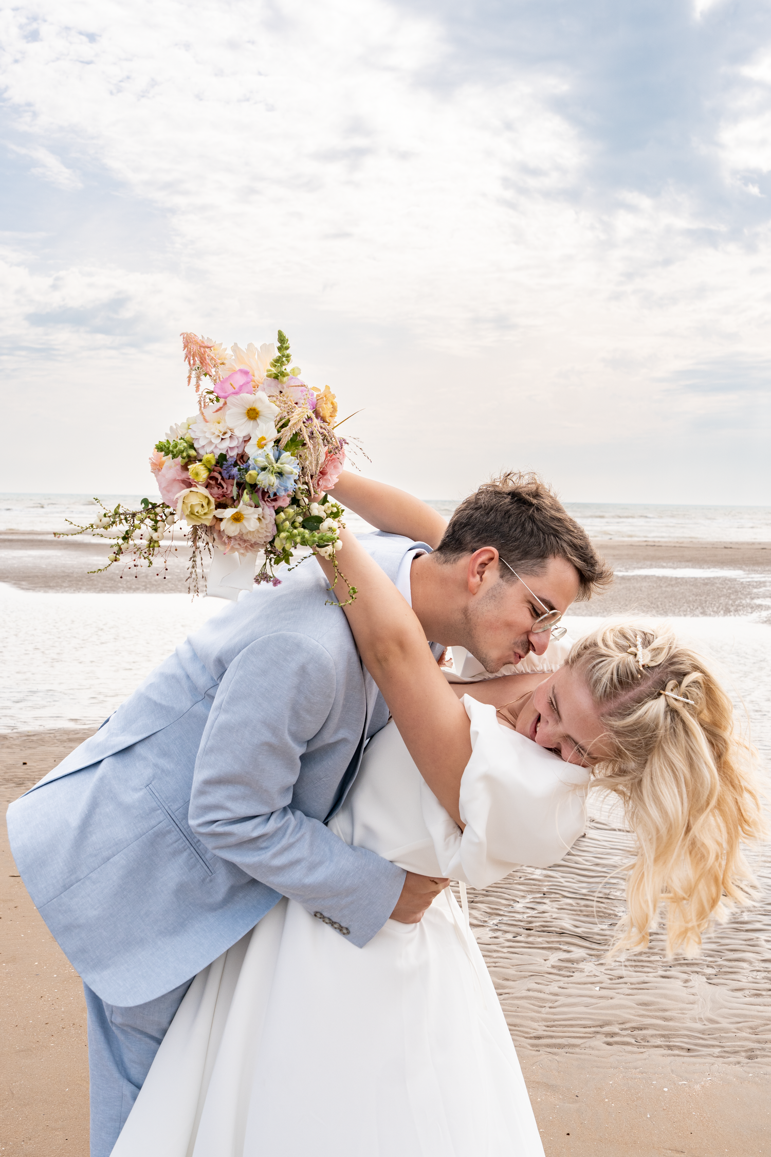 Groom in light blue suit kissing bride in white dress on beach, holding colorful bouquet.
