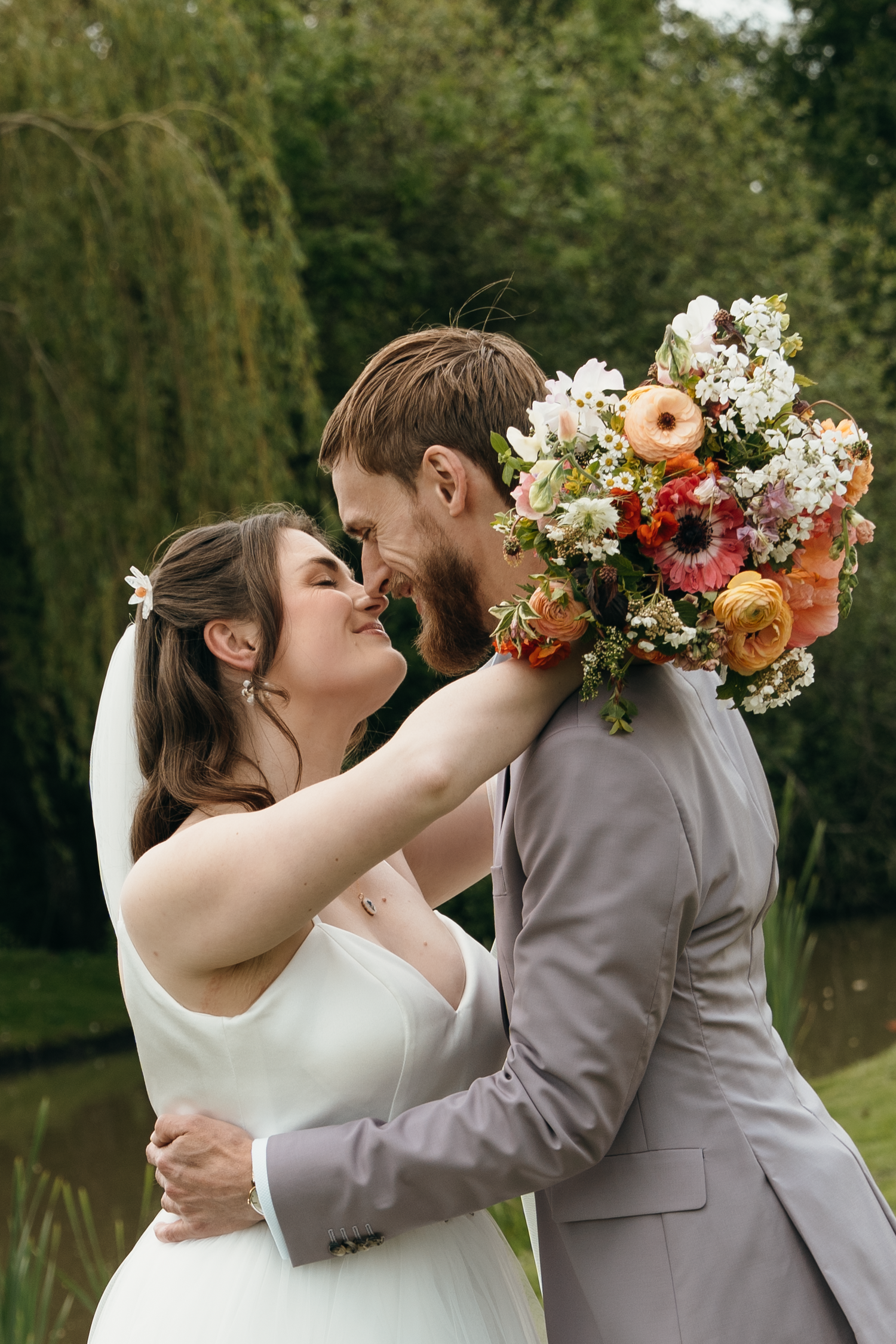 Bride and groom share a romantic moment, holding a colorful bouquet, surrounded by lush greenery outdoors.