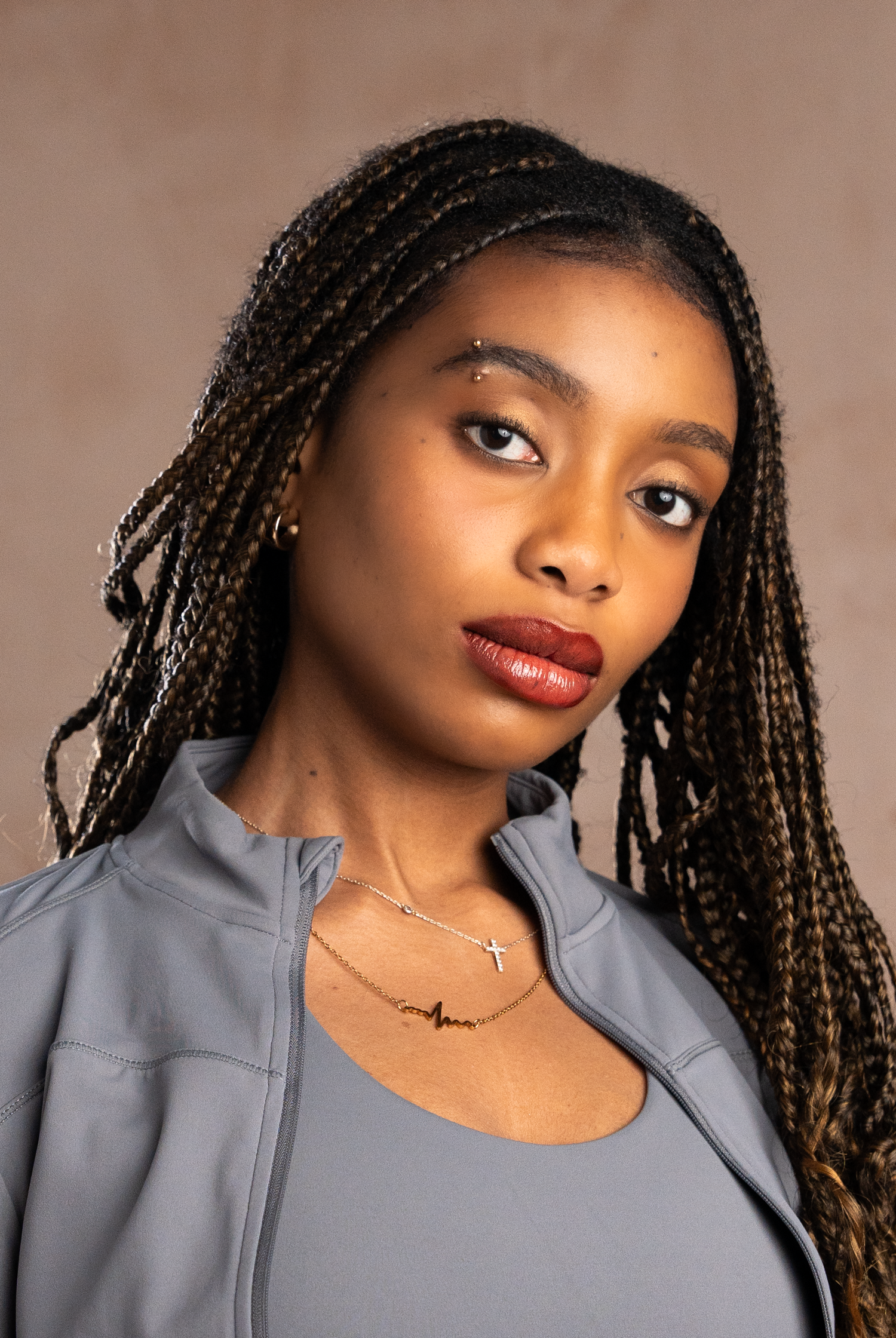 Young woman with braids, nose piercing, red lips, wearing grey zip-up jacket and delicate cross necklace.