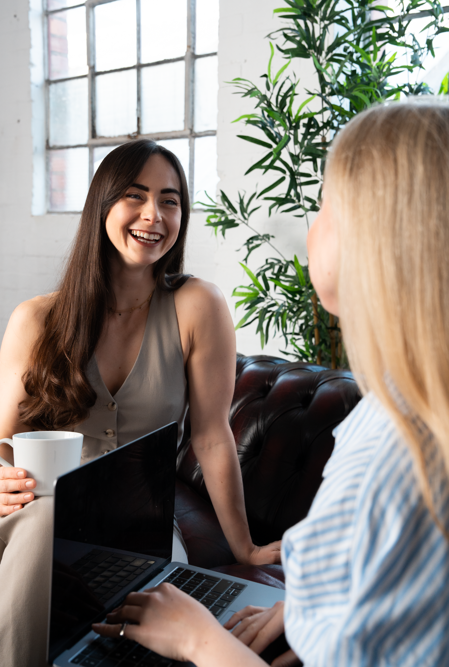 Two women smile and chat over laptops and coffee in a bright, plant-filled modern office lounge setting.