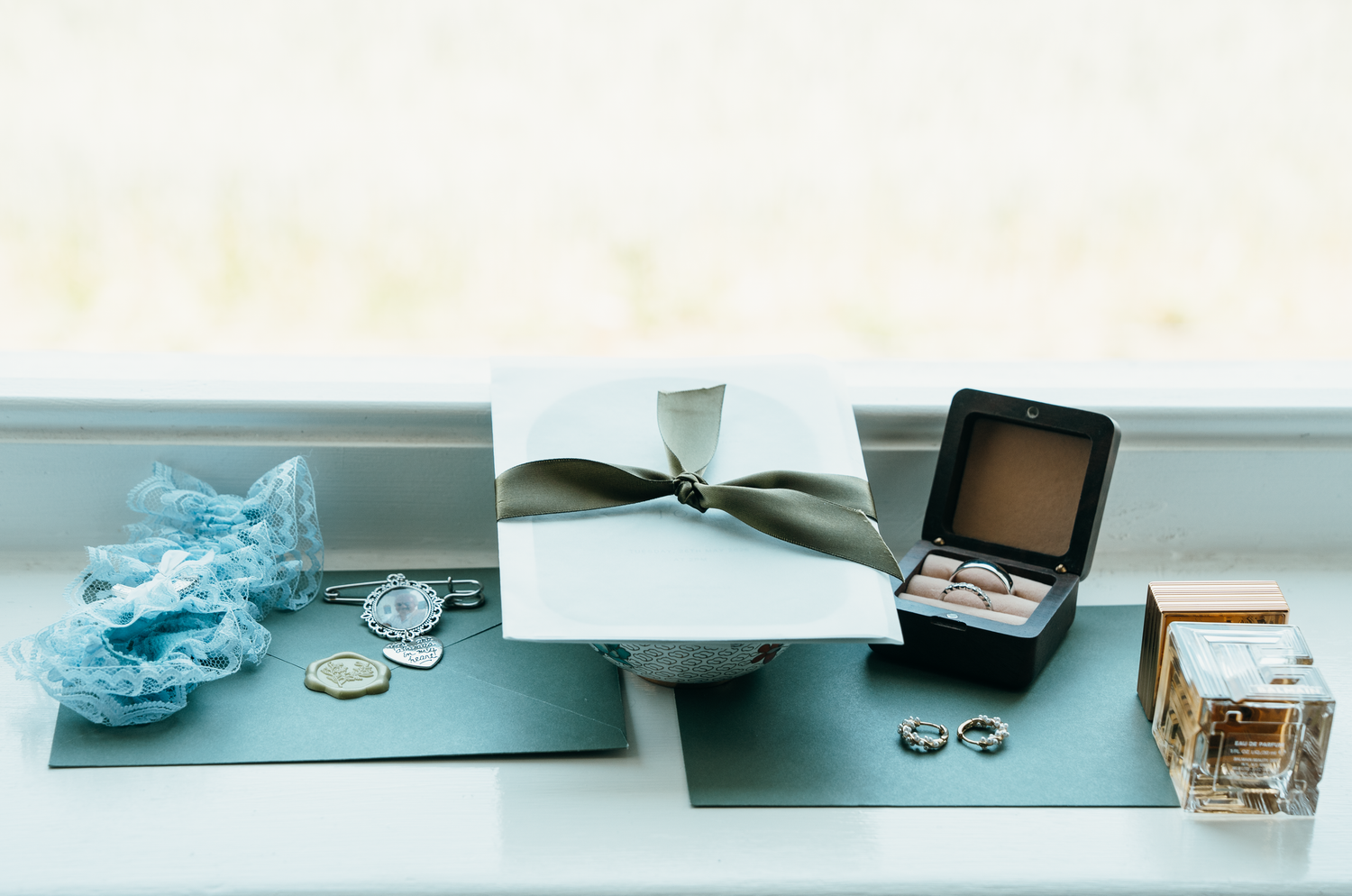 Wedding flat lay with ring box, rings, gift box, blue lace garter, coins, and perfume on a windowsill.