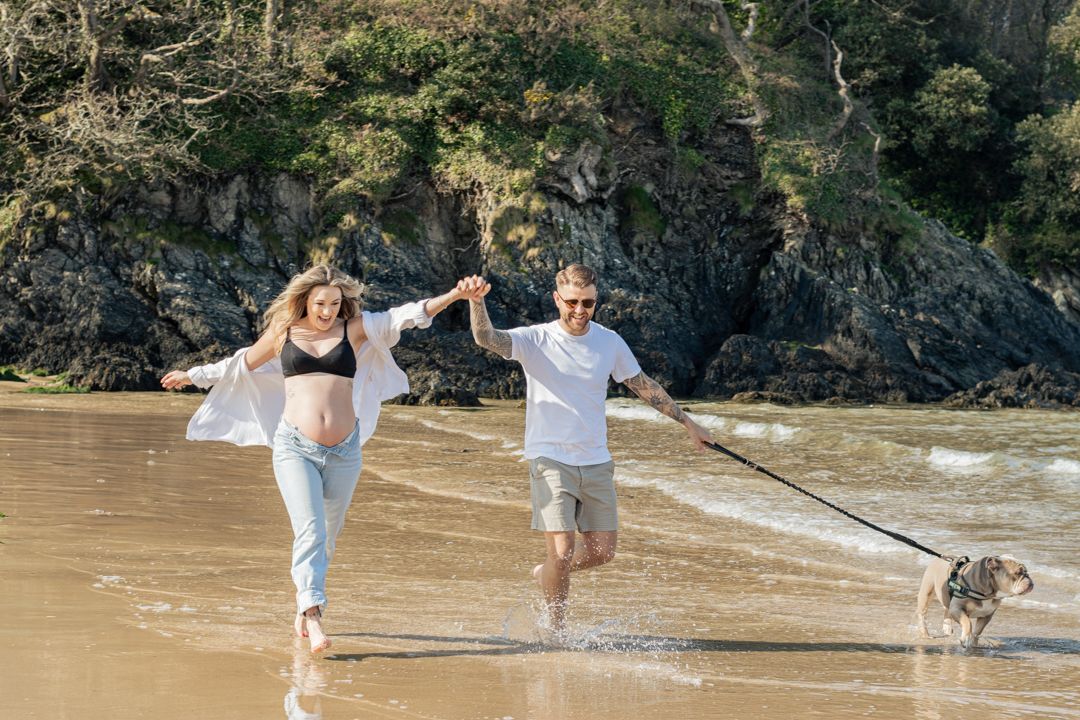 Joyful pregnant couple running through ocean waves with bulldog on sunny beach.