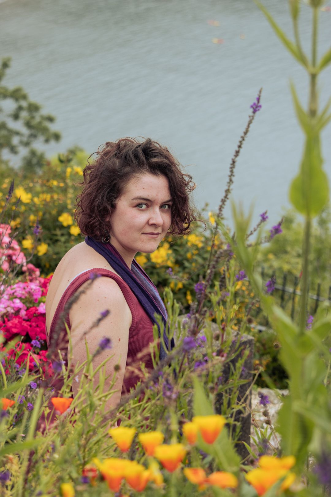 Young woman in burgundy dress sits among colorful wildflowers with turquoise water in background.