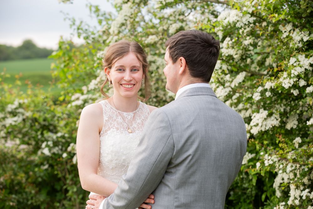 A smiling bride in lace dress holds hands with groom near blooming white flowers outdoors.