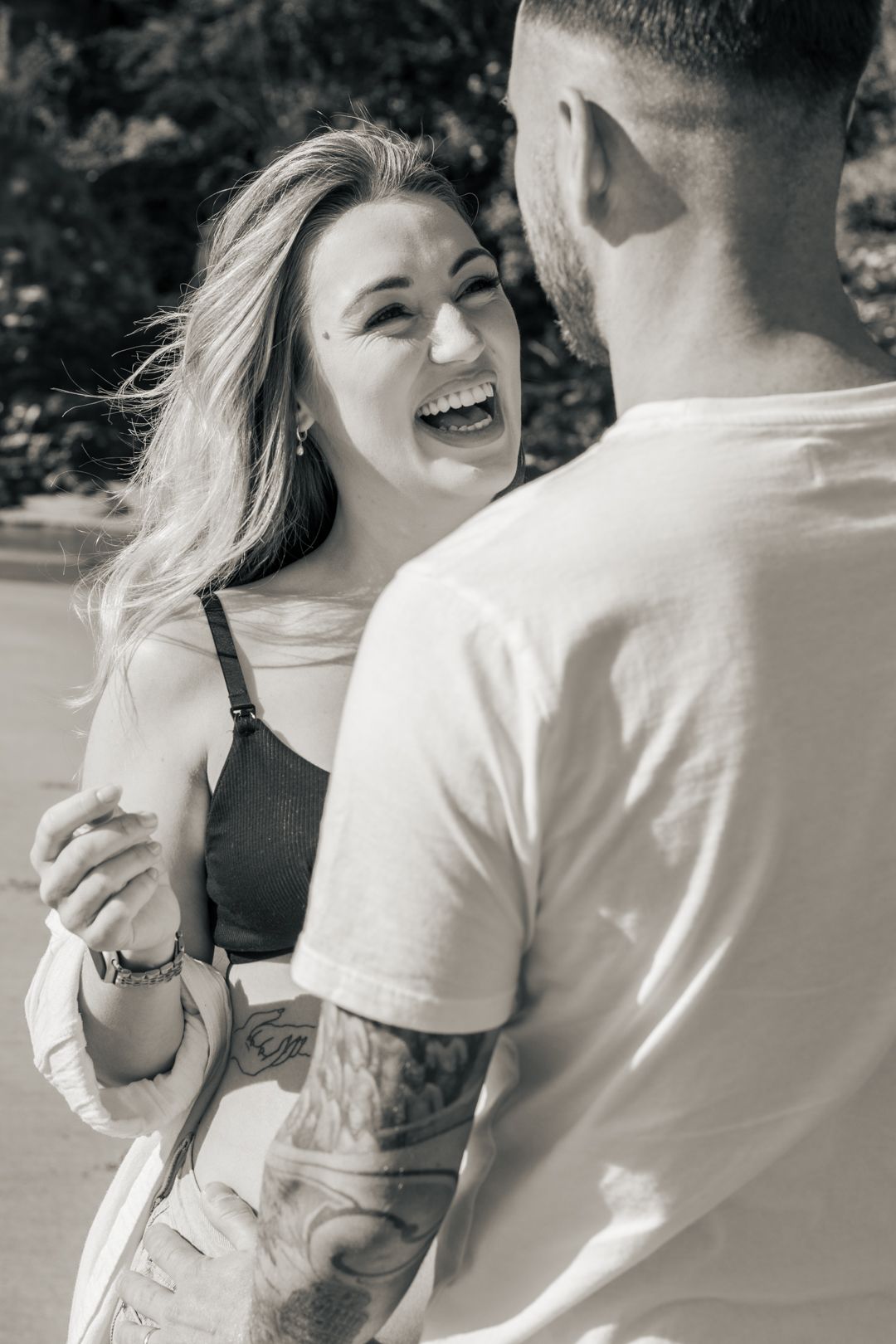 Black and white photo of laughing pregnant woman dancing with tattooed partner outdoors.