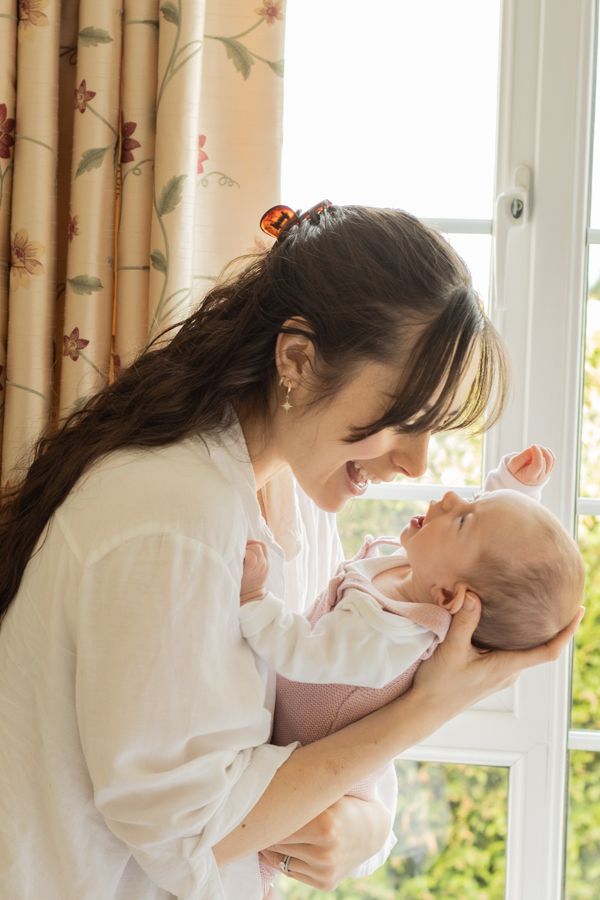 Happy mother smiling and holding newborn baby near bright window indoors.