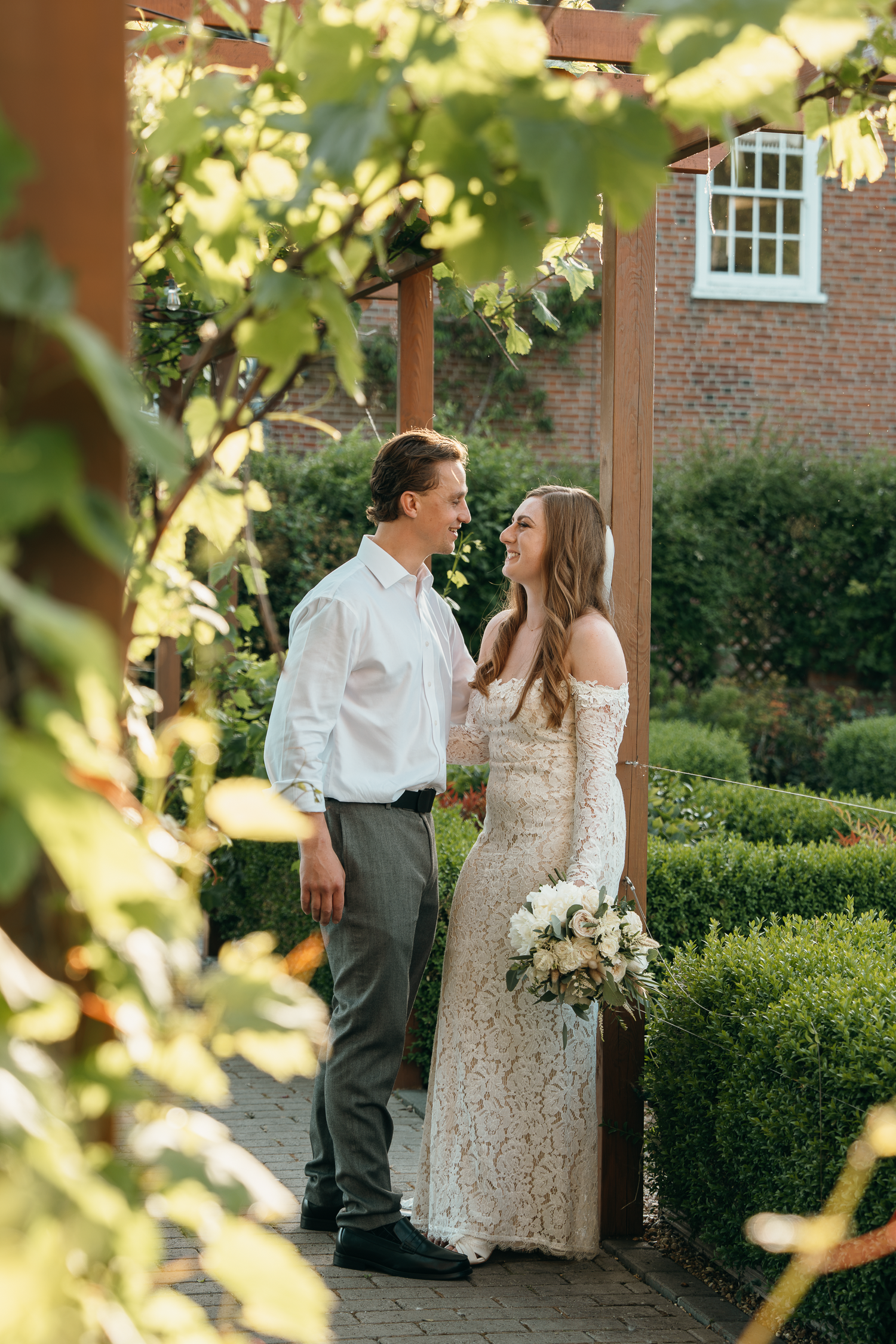 A smiling bride in lace gown holding white bouquet stands with groom in white shirt in a lush garden setting.