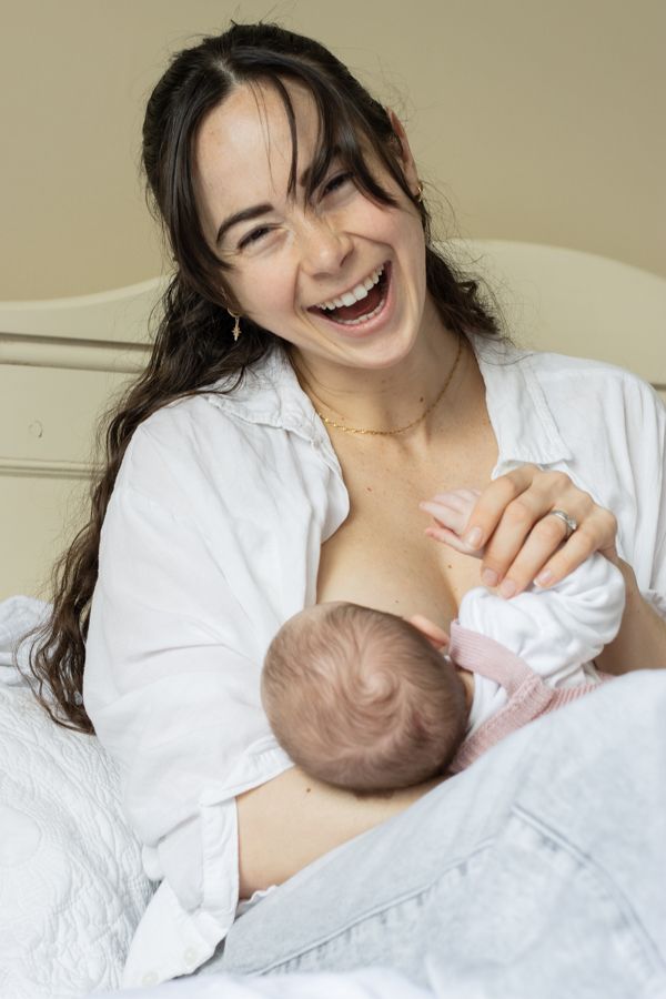 A joyful mother laughing while breastfeeding her newborn baby on a bed.