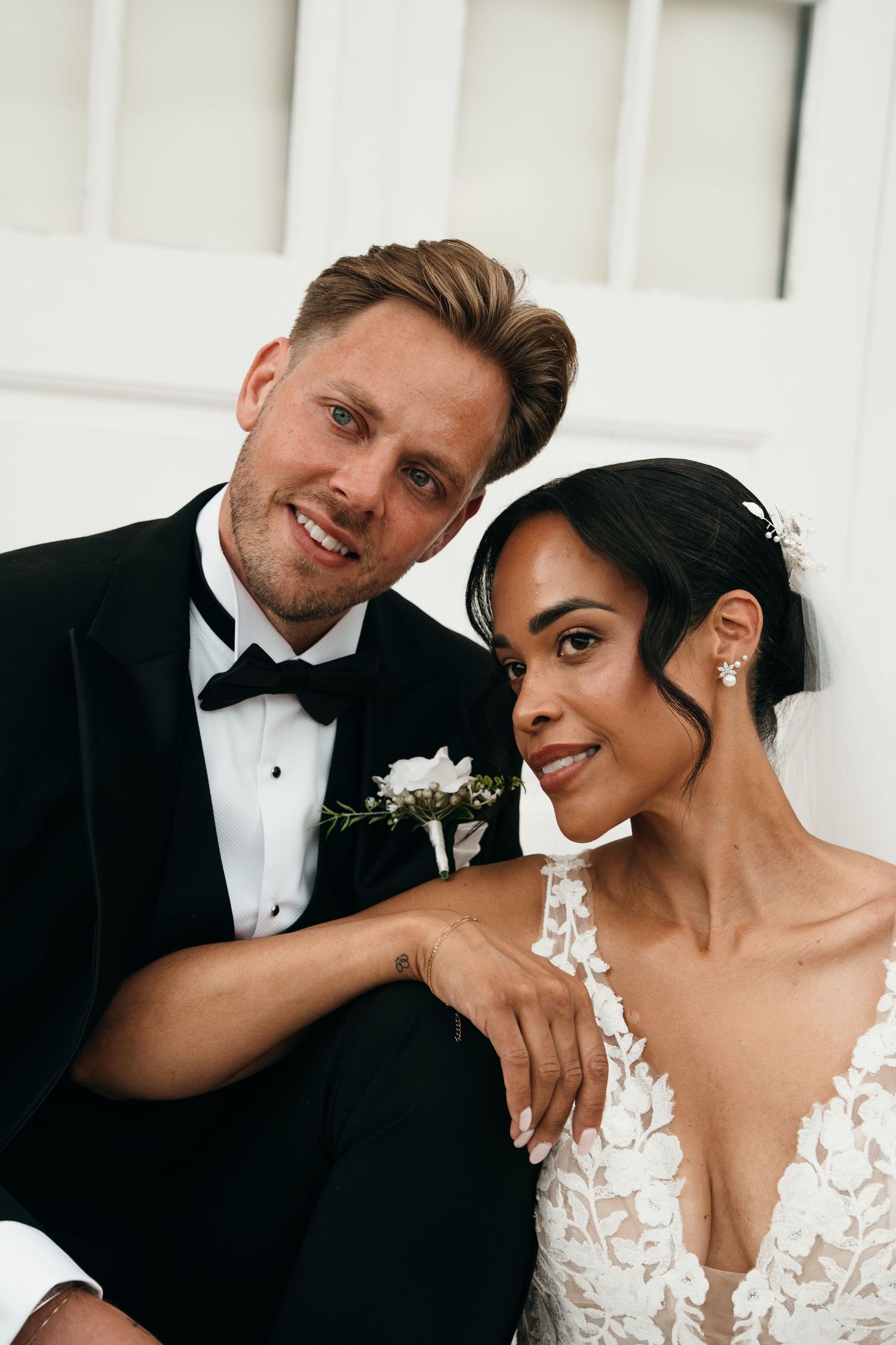 A smiling groom in a black tuxedo and bride in lace wedding dress pose together for a romantic portrait photo.