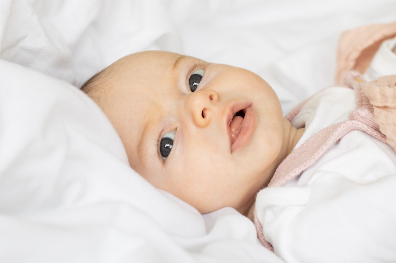 A wide-eyed newborn baby lying on soft white bedding with mouth slightly open.