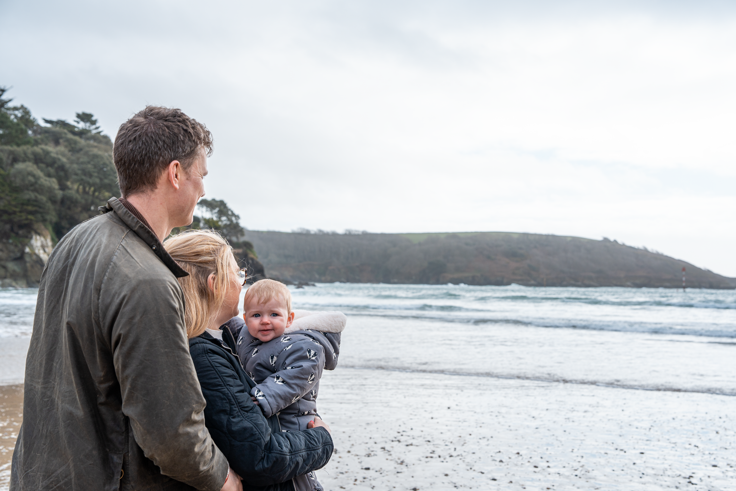Family with baby gazing at ocean waves on a cloudy beach day.