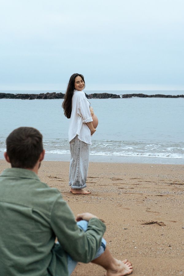 Pregnant woman in white shirt smiling over shoulder at partner sitting on sandy beach by ocean.