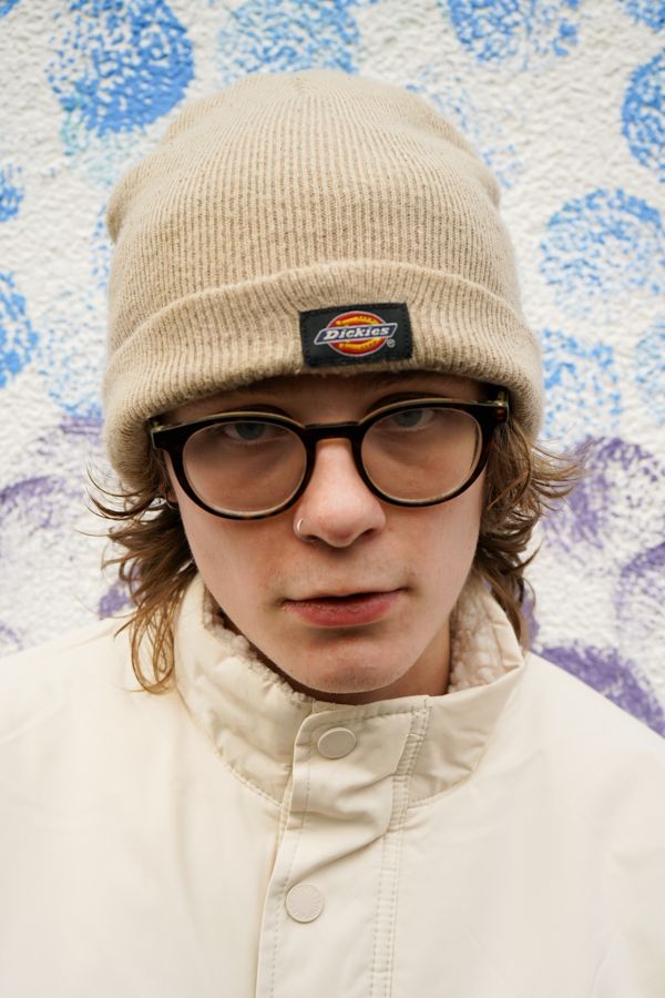 Close-up portrait of young person wearing Dickies beanie and glasses against blue dotted wall.