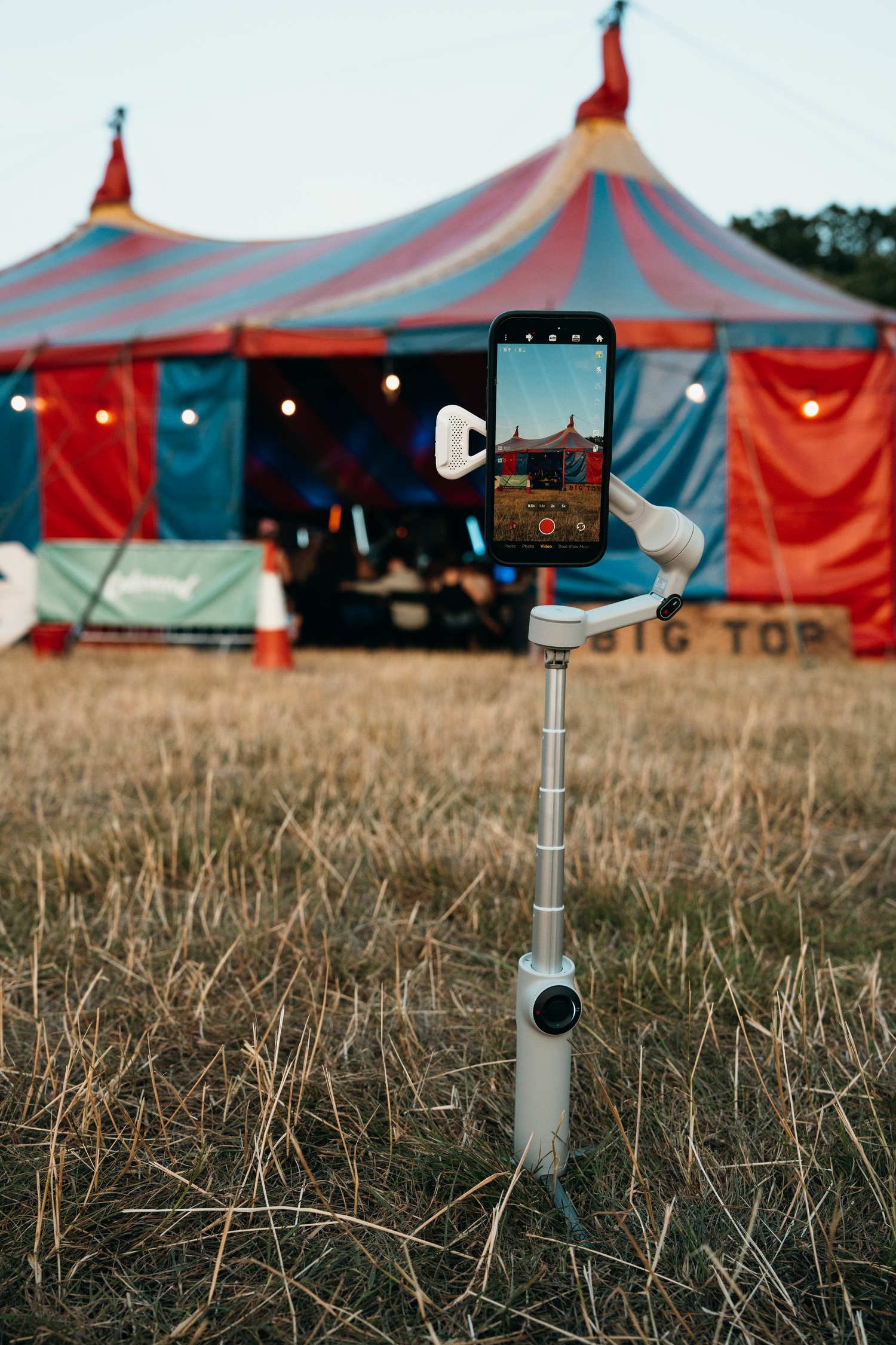 Smartphone on a gimbal stabilizer filming a colorful circus big top tent at a festival in a dry grass field.
