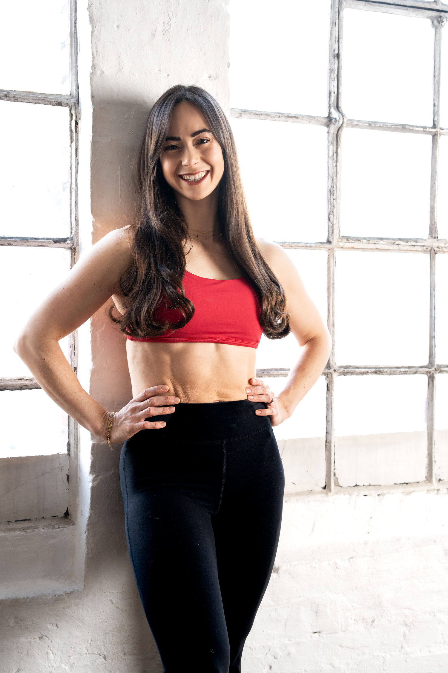 A smiling athletic woman in a red crop top and black leggings poses in a bright studio.