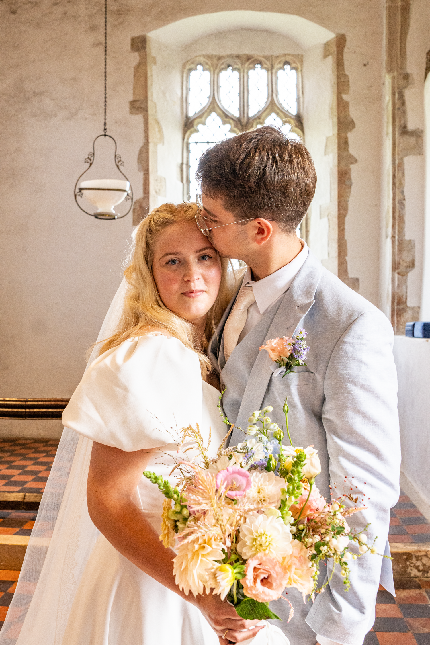 Bride and groom posing in a rustic church interior, groom kissing bride's head while she holds a pastel bouquet.