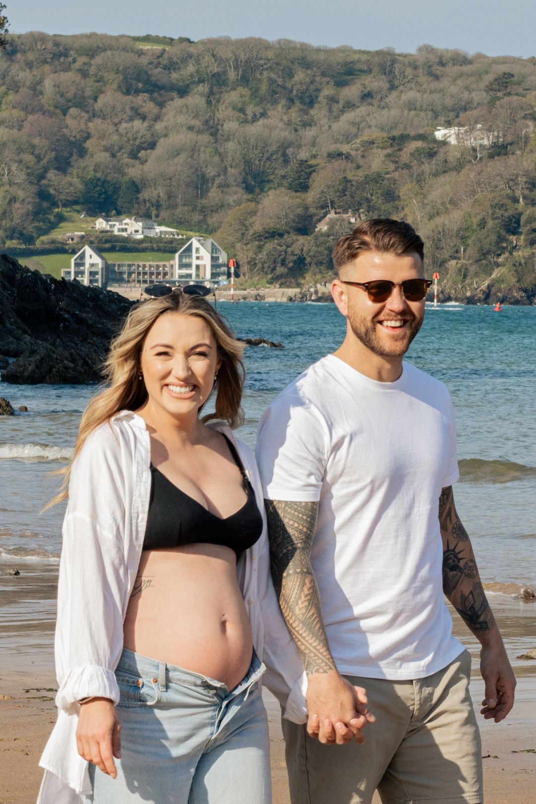 Smiling pregnant couple holding hands on a scenic beach with rocky coastline and blue water behind them.