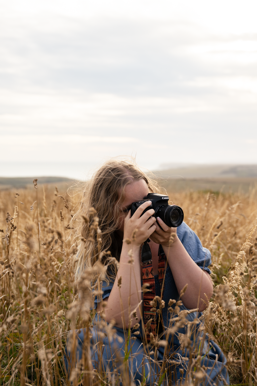Blonde woman photographing through camera lens in tall golden wheat field.