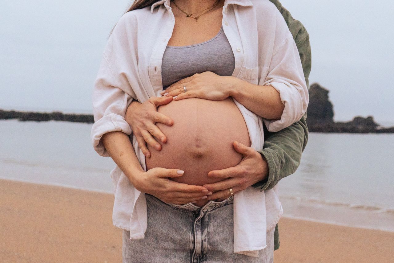Close-up of pregnant belly with couple holding bump together on beach.