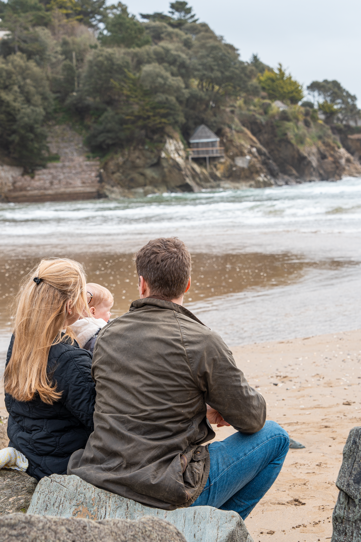 Family of three sitting on rocks watching waves on a beach.