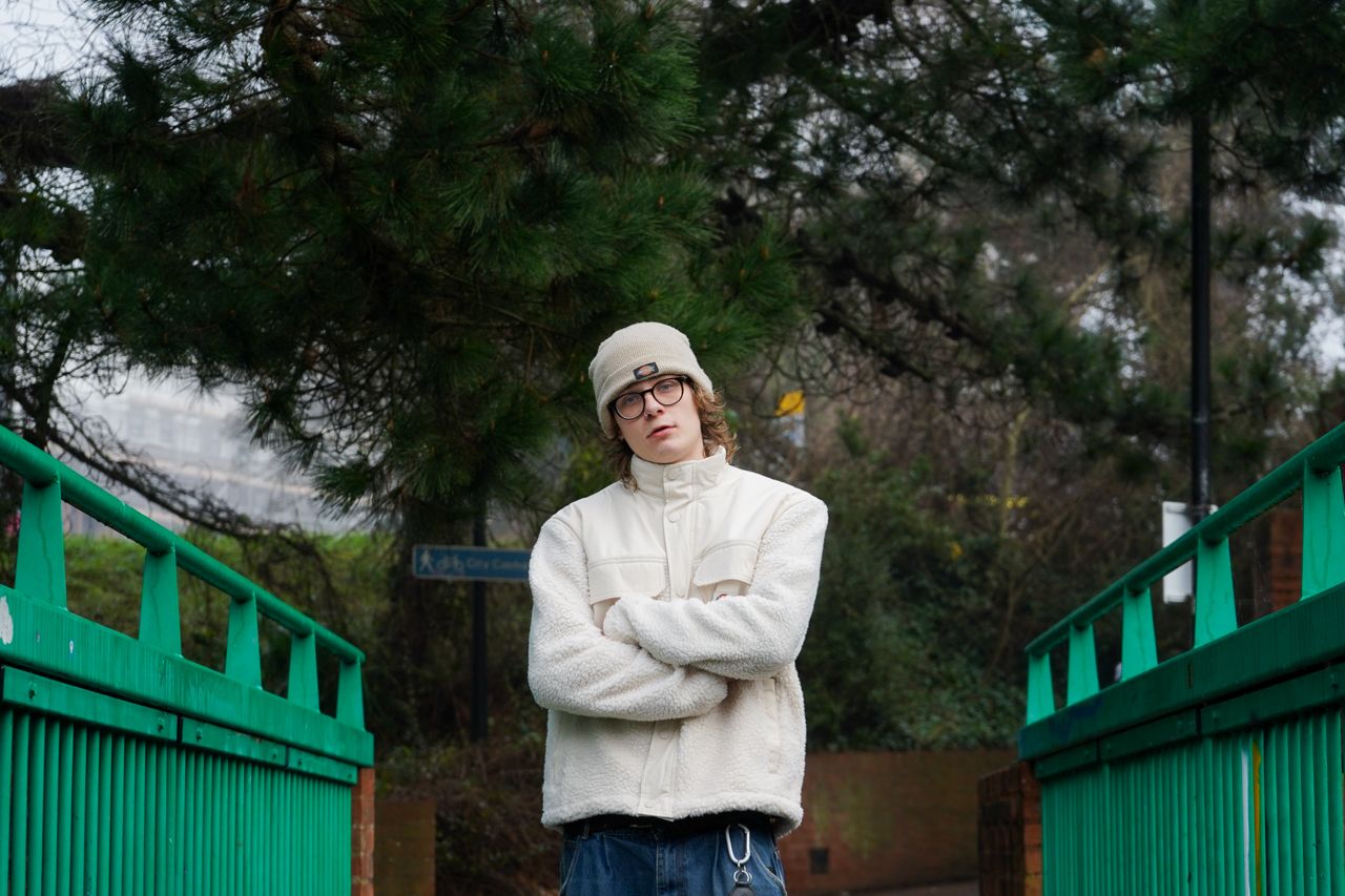 Person in white fleece jacket and beanie standing arms crossed on green bridge outdoors.