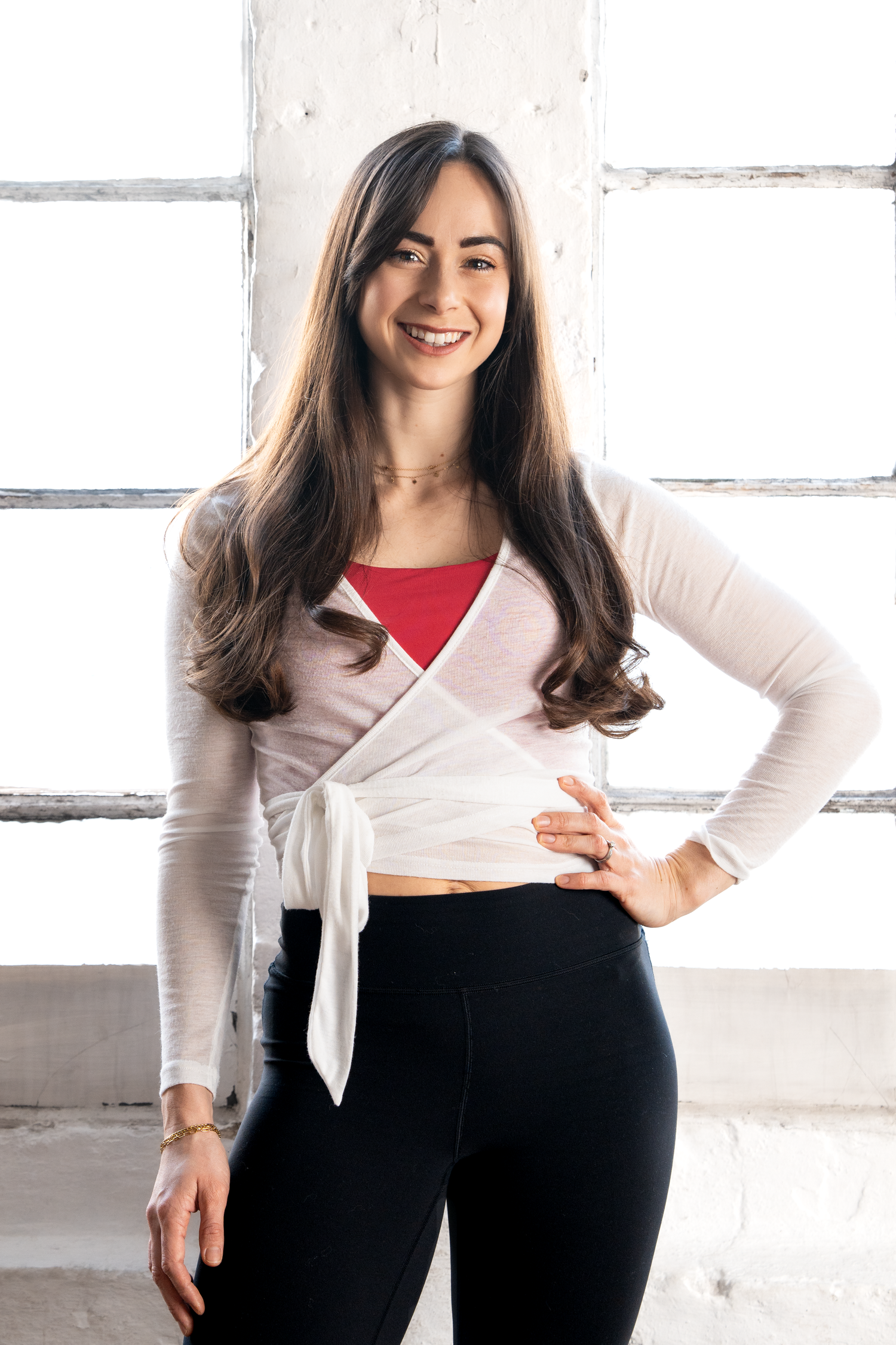 Smiling young woman with long brown hair wearing a white wrap top and black leggings posing in a bright studio.