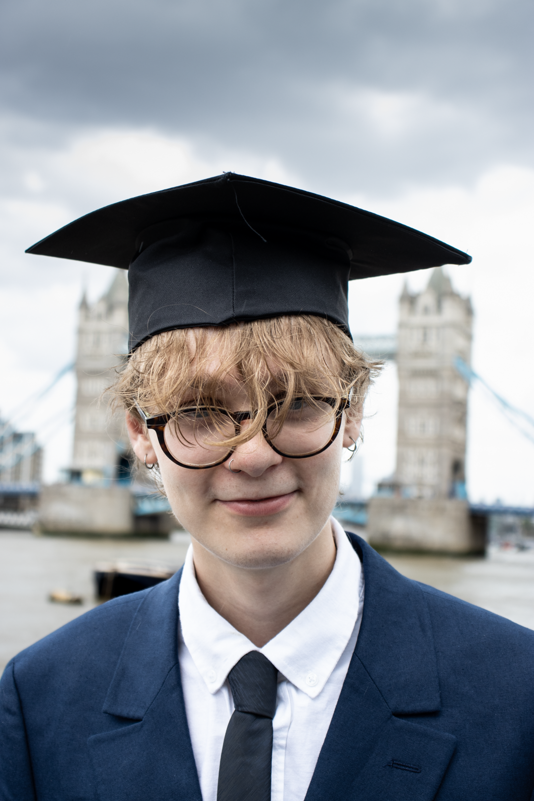 A young graduate wearing glasses and a graduation cap smiles with Tower Bridge in the background.