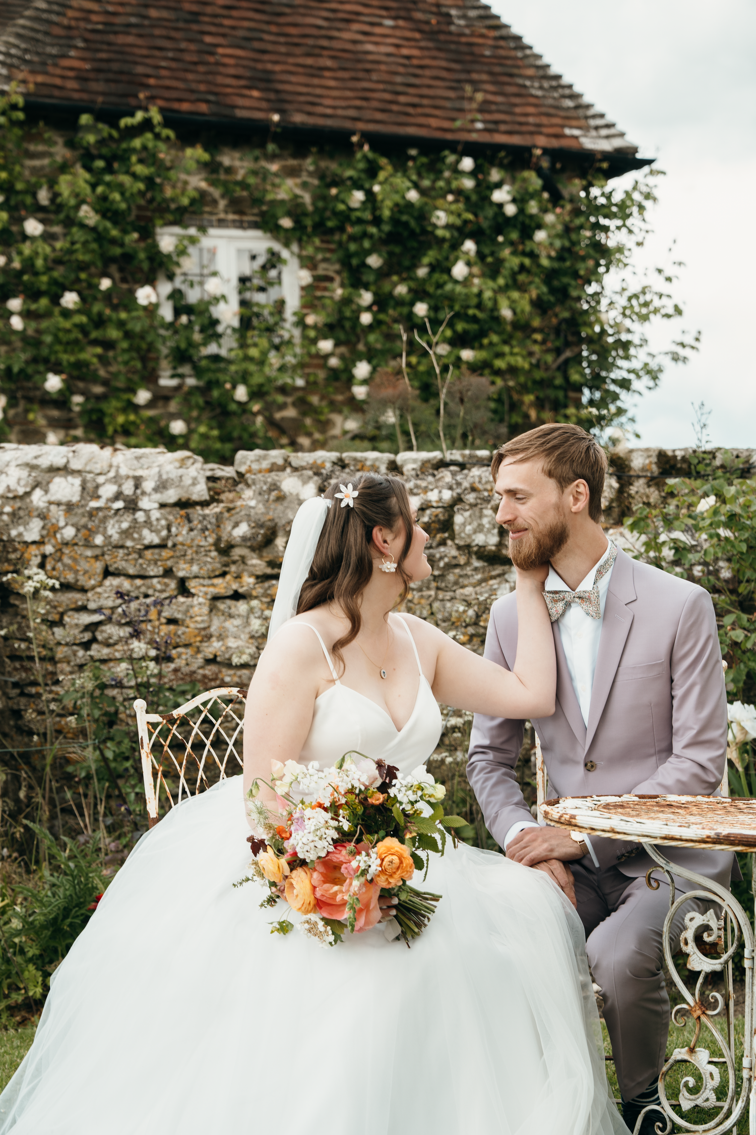 Bride and groom sit at outdoor table with colorful bouquet, smiling at each other before ivy-covered stone building.