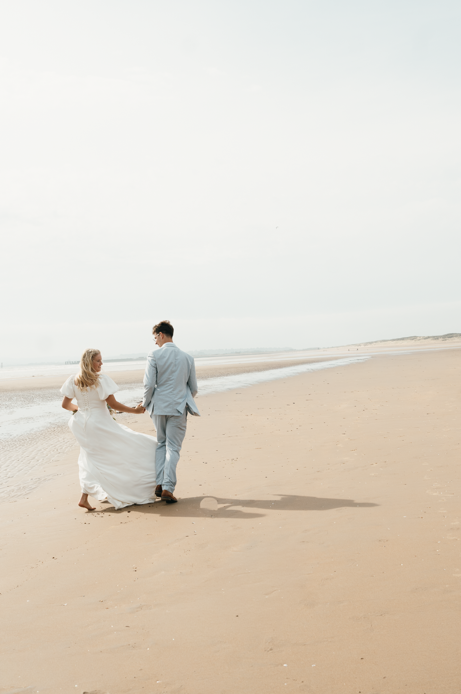A bride and groom walk hand-in-hand along a sandy beach, her white gown flowing in the breeze.